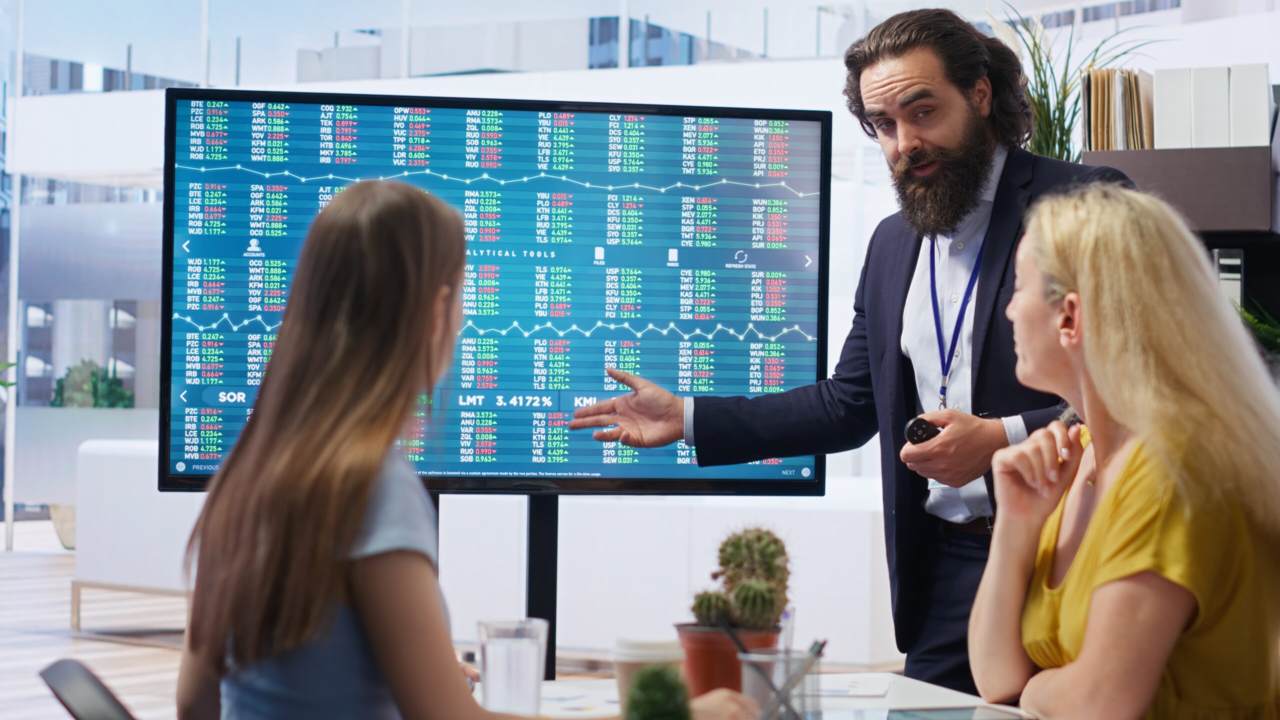 A financial analyst explaining time series analysis to two women in an office, with a large screen displaying stock market data and charts in the background.